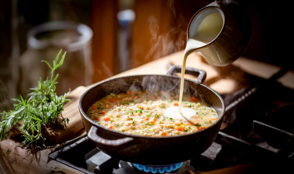 Cremige Suppe köchelt in einem Topf auf dem Herd, während Sahne hineingegossen wird; frische Kräuter liegen daneben.